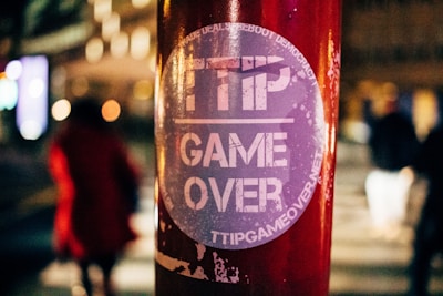 A round sticker with the text 'TTIP GAME OVER' is placed on a red pole. The background is blurred with silhouettes of people walking, indicating a nighttime setting with bright city lights.