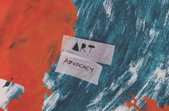 A textured abstract background with bold strokes of orange and dark teal. Two pieces of paper with the words 'Art' and 'Advocacy' written on them are prominently placed in the center.