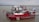 A red fire rescue boat with various equipment and hoses is docked on a calm body of water. The boat has flags on either side and several people in uniform are aboard. The background shows a cloudy sky and a distant tree-lined shoreline.