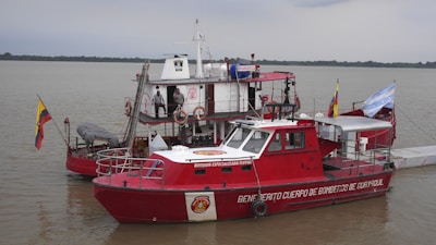 A red fire rescue boat with various equipment and hoses is docked on a calm body of water. The boat has flags on either side and several people in uniform are aboard. The background shows a cloudy sky and a distant tree-lined shoreline.