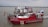 A red fire rescue boat with various equipment and hoses is docked on a calm body of water. The boat has flags on either side and several people in uniform are aboard. The background shows a cloudy sky and a distant tree-lined shoreline.