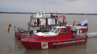 A red fire rescue boat with various equipment and hoses is docked on a calm body of water. The boat has flags on either side and several people in uniform are aboard. The background shows a cloudy sky and a distant tree-lined shoreline.