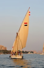 A traditional sailboat, known as a felucca, is sailing on a calm body of water. The large sail, adorned with the colors of a national flag, catches the breeze as the sun sets, casting a warm glow over the scene. In the background, a cityscape with buildings and greenery lines the horizon.