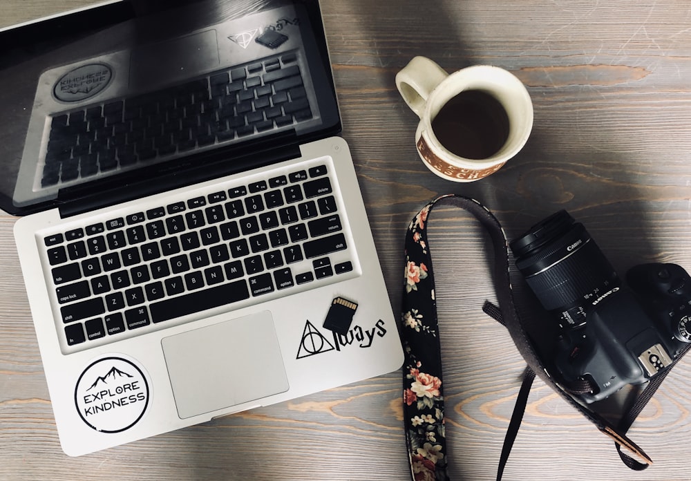 computador portátil preto e cinza ao lado da câmera DSLR preta e caneca de cerâmica branca