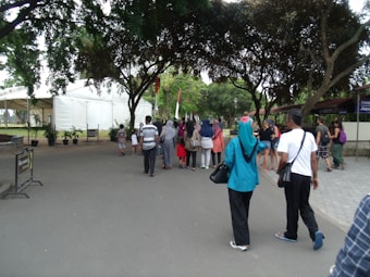A group of people walking along a wide pathway lined with trees on both sides. To the left, a large white tent and some plants are visible, while the right side shows buildings partially covered by trees. The pathway is surrounded by greenery and the people appear diverse in clothing and style, suggesting a casual outdoor event or gathering.