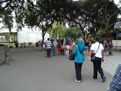 A group of people walking along a wide pathway lined with trees on both sides. To the left, a large white tent and some plants are visible, while the right side shows buildings partially covered by trees. The pathway is surrounded by greenery and the people appear diverse in clothing and style, suggesting a casual outdoor event or gathering.