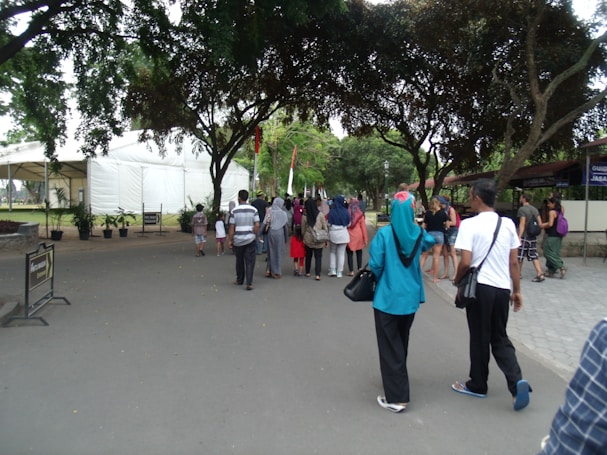 A group of people walking along a wide pathway lined with trees on both sides. To the left, a large white tent and some plants are visible, while the right side shows buildings partially covered by trees. The pathway is surrounded by greenery and the people appear diverse in clothing and style, suggesting a casual outdoor event or gathering.
