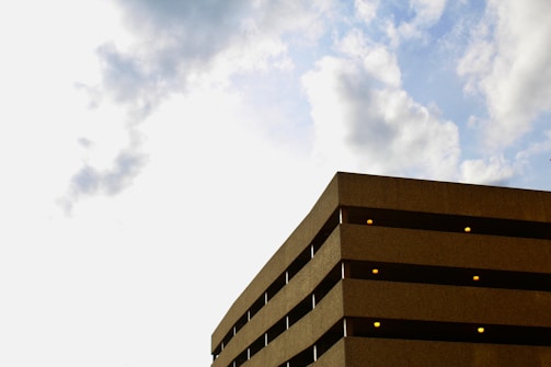A concrete parking structure with multiple levels is visible against a backdrop of a partly cloudy sky. The building features rows of small, evenly spaced lights along each level, and the clouds appear soft and fluffy.
