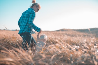 woman holding toddler waling near brown grass during daytime