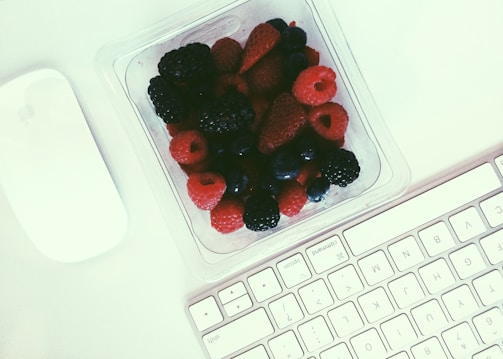A clear plastic container filled with an assortment of fresh berries, including raspberries, blackberries, and blueberries. The container is placed on a white surface, next to a wireless mouse and a computer keyboard, suggesting an office or work environment.