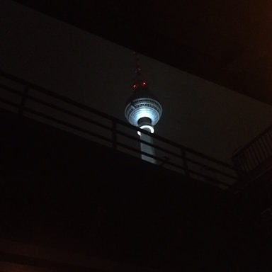 A brightly lit telecommunications tower is seen against a dark background, partially obscured by a silhouetted bridge or overpass. The tower features an illuminated sphere near the top and several red lights along its length.