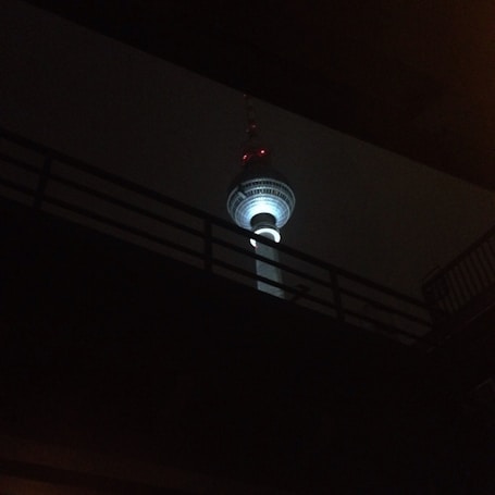 A brightly lit telecommunications tower is seen against a dark background, partially obscured by a silhouetted bridge or overpass. The tower features an illuminated sphere near the top and several red lights along its length.