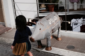A child with dark hair in a navy top and brown skirt walks past a metal sculpture resembling an animal with the words 'Pigsty Used Clothing' written on its side. The setting is an outdoor area near a shop with clothes displayed inside.