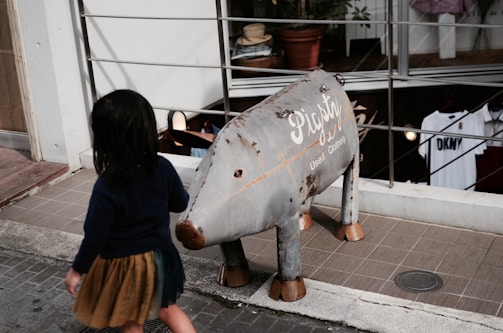 A child with dark hair in a navy top and brown skirt walks past a metal sculpture resembling an animal with the words 'Pigsty Used Clothing' written on its side. The setting is an outdoor area near a shop with clothes displayed inside.