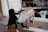 A child with dark hair in a navy top and brown skirt walks past a metal sculpture resembling an animal with the words 'Pigsty Used Clothing' written on its side. The setting is an outdoor area near a shop with clothes displayed inside.
