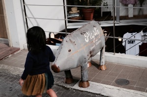 A child with dark hair in a navy top and brown skirt walks past a metal sculpture resembling an animal with the words 'Pigsty Used Clothing' written on its side. The setting is an outdoor area near a shop with clothes displayed inside.