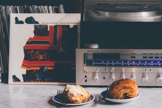 A vintage stereo receiver and turntable are placed on a table next to a vinyl record with a colorful abstract cover. In front of the stereo system, there are two plates, one holding a croissant, and the other a pastry with icing. The background shows a collection of vinyl records.