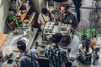 A bustling coffee shop scene with people gathered around a coffee machine. The baristas are preparing drinks, interacting with customers. Various coffee-making equipment and cups are neatly arranged on the counter. The atmosphere appears inviting and lively, with plants and shelves stocked with different products adding to the cozy ambiance.