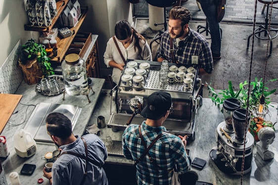 A bustling coffee shop scene with people gathered around a coffee machine. The baristas are preparing drinks, interacting with customers. Various coffee-making equipment and cups are neatly arranged on the counter. The atmosphere appears inviting and lively, with plants and shelves stocked with different products adding to the cozy ambiance.