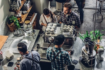 A bustling coffee shop scene with people gathered around a coffee machine. The baristas are preparing drinks, interacting with customers. Various coffee-making equipment and cups are neatly arranged on the counter. The atmosphere appears inviting and lively, with plants and shelves stocked with different products adding to the cozy ambiance.
