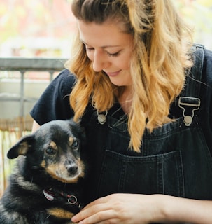 A woman with blonde hair smiles affectionately while holding a small black and brown dog. She wears a black shirt with denim overalls and is seated outdoors with a blurred background.