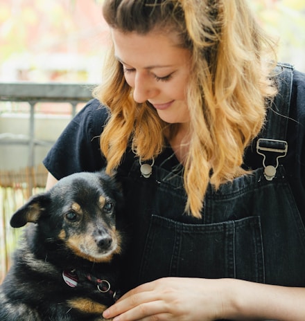 A woman with blonde hair smiles affectionately while holding a small black and brown dog. She wears a black shirt with denim overalls and is seated outdoors with a blurred background.