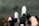 A pair of worn sneakers and black shoes are visible against a gray background. One person wears white sneakers with red and black accents, while another has black shoes. Both individuals have ripped black jeans, adding an edgy style to the scene.