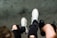 A pair of worn sneakers and black shoes are visible against a gray background. One person wears white sneakers with red and black accents, while another has black shoes. Both individuals have ripped black jeans, adding an edgy style to the scene.