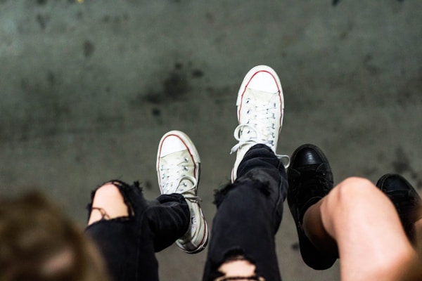 A pair of worn sneakers and black shoes are visible against a gray background. One person wears white sneakers with red and black accents, while another has black shoes. Both individuals have ripped black jeans, adding an edgy style to the scene.