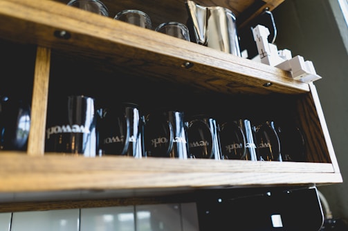 A collection of clean, modern mugs arranged neatly on a light wooden shelf, bathed in natural daylight.