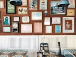 A wall decorated with an eclectic mix of colorful framed posters and artwork, each with different themes and messages. The frames vary in size and style, adding a vibrant and artistic flair. Below the wall, there is a table with a patterned surface, a black chair, and a bottle. A guitar leans against the wall, contributing to the creative ambiance.