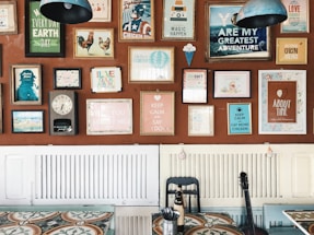 A wall decorated with an eclectic mix of colorful framed posters and artwork, each with different themes and messages. The frames vary in size and style, adding a vibrant and artistic flair. Below the wall, there is a table with a patterned surface, a black chair, and a bottle. A guitar leans against the wall, contributing to the creative ambiance.