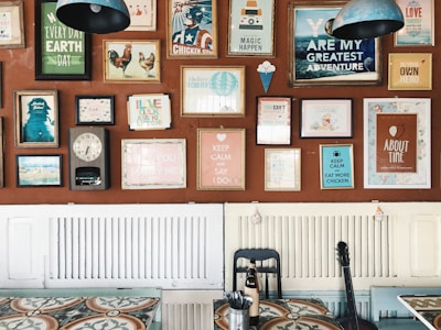 A wall decorated with an eclectic mix of colorful framed posters and artwork, each with different themes and messages. The frames vary in size and style, adding a vibrant and artistic flair. Below the wall, there is a table with a patterned surface, a black chair, and a bottle. A guitar leans against the wall, contributing to the creative ambiance.