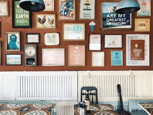 A wall decorated with an eclectic mix of colorful framed posters and artwork, each with different themes and messages. The frames vary in size and style, adding a vibrant and artistic flair. Below the wall, there is a table with a patterned surface, a black chair, and a bottle. A guitar leans against the wall, contributing to the creative ambiance.