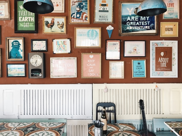 A wall decorated with an eclectic mix of colorful framed posters and artwork, each with different themes and messages. The frames vary in size and style, adding a vibrant and artistic flair. Below the wall, there is a table with a patterned surface, a black chair, and a bottle. A guitar leans against the wall, contributing to the creative ambiance.
