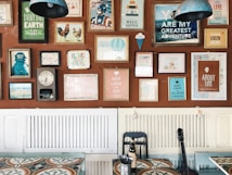 A wall decorated with an eclectic mix of colorful framed posters and artwork, each with different themes and messages. The frames vary in size and style, adding a vibrant and artistic flair. Below the wall, there is a table with a patterned surface, a black chair, and a bottle. A guitar leans against the wall, contributing to the creative ambiance.