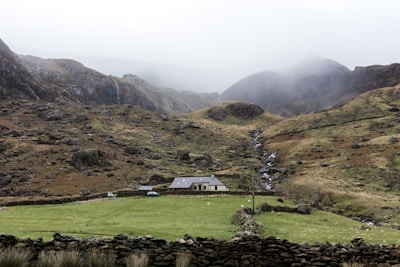 A rural landscape featuring a small house with a sloped roof situated in a lush green field. The background is dominated by rugged, mist-covered hills and mountains. A narrow waterfall flows down the rocky hillside, adding a sense of movement to the tranquil scene. Stone walls divide the property, creating a picturesque and serene atmosphere.