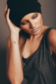 A fashionably dressed woman poses with her head tilted to the side, gently holding a black beanie. Her eyes are closed, and she has metallic eye makeup and lip color. A tattoo is visible on her forearm, and she wears a sleeveless gray top.