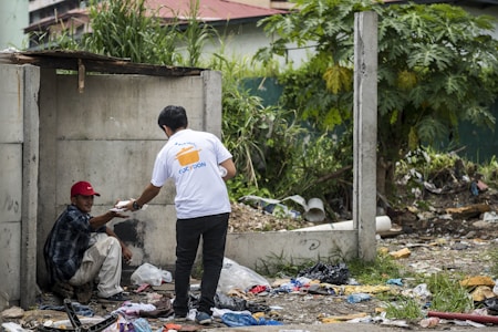 A man wearing a red hat sits on a concrete ledge amidst a pile of trash and debris while another person wearing jeans and a white t-shirt hands him a small package. The area is surrounded by green vegetation and an unfinished concrete structure, suggesting a setting of urban poverty.