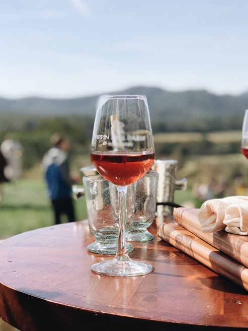 A glass of red wine sits elegantly on a wooden table outdoors. Two empty glasses and a folded napkin are nearby, with a vineyard and rolling hills in the blurred background under a clear blue sky.
