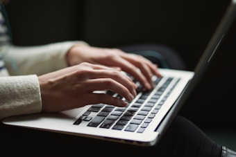 Hands are typing on a laptop keyboard. The left sleeve of the person appears to be rolled slightly upwards, and the background is dark and out of focus.