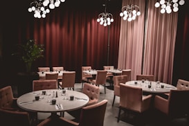 A stylish and elegant restaurant interior featuring round tables set with glassware and napkins, surrounded by plush, upholstered chairs. Soft, ambient lighting cascades from modern chandeliers, casting a warm glow on the space. Rich, floor-to-ceiling maroon curtains add a touch of luxury, while a potted green plant adds a natural element to the scene.