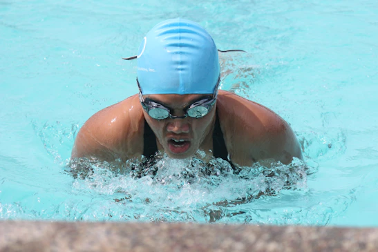 Close-up of a determined athlete in a sleek swim cap and goggles, poised in deep blue water with sunlight reflections.