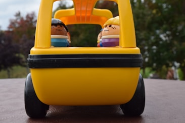 A close-up of a toy bus with small figurines inside. The bus is predominantly yellow with a black stripe and visible wheels. In the background, there is some nature with greenery and trees.
