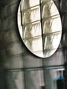 A circular mirror mounted on a wall made of textured glass blocks. The mirror reflects the glass blocks, creating a pattern of geometric shapes and lines, with a subtle play of light and shadow.