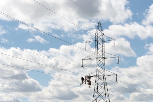 A large metal electricity pylon stands against a partly cloudy blue sky. Several power lines stretch horizontally across the view. Two workers are suspended mid-air on a platform, possibly performing maintenance or installation tasks on the pylon.