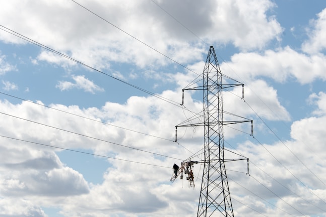 A large metal electricity pylon stands against a partly cloudy blue sky. Several power lines stretch horizontally across the view. Two workers are suspended mid-air on a platform, possibly performing maintenance or installation tasks on the pylon.