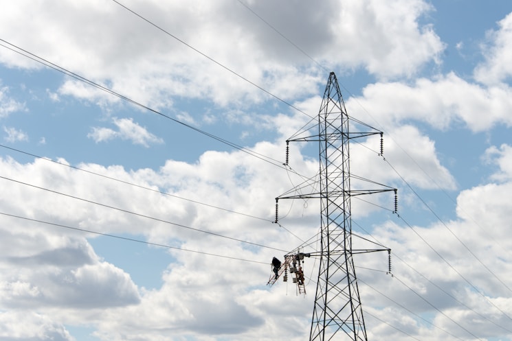 A large metal electricity pylon stands against a partly cloudy blue sky. Several power lines stretch horizontally across the view. Two workers are suspended mid-air on a platform, possibly performing maintenance or installation tasks on the pylon.