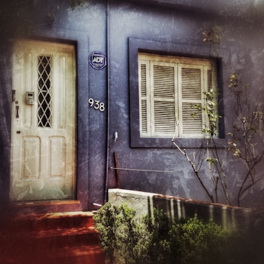 A weathered blue facade of a building features a white door with a diamond-patterned window and an adjacent number '938'. A security company sign is mounted near the door. Beside the door, there is a window with closed shutters. Sparse foliage and some bare branches can be seen in front of the building.