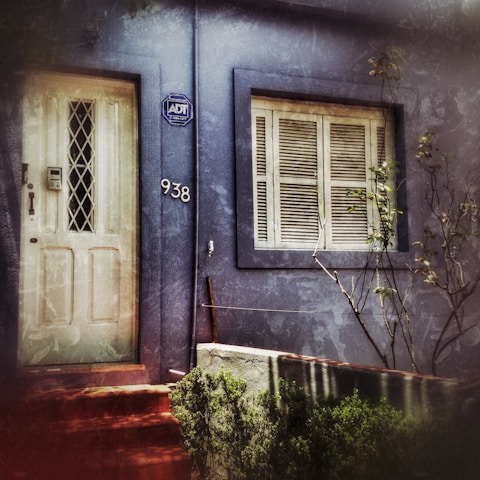 A weathered blue facade of a building features a white door with a diamond-patterned window and an adjacent number '938'. A security company sign is mounted near the door. Beside the door, there is a window with closed shutters. Sparse foliage and some bare branches can be seen in front of the building.